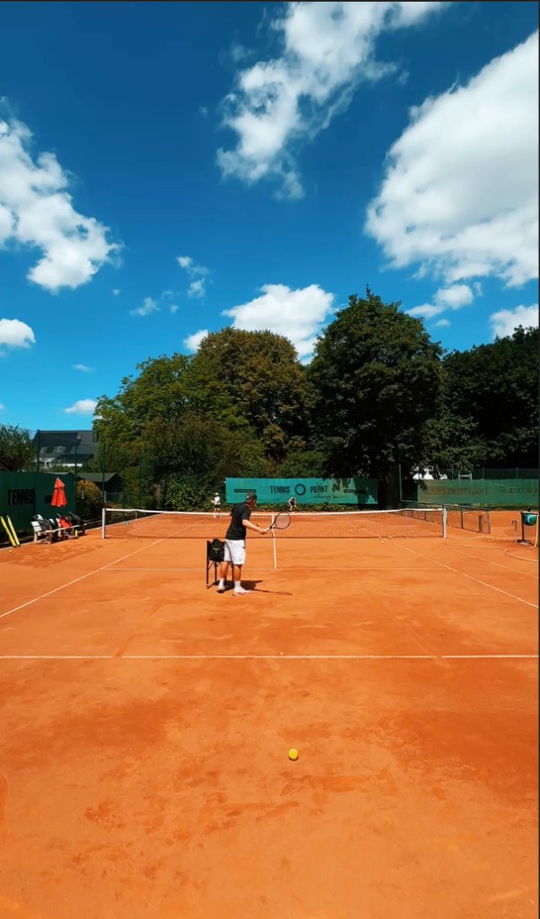 Trainer mit Schläger und gefülltem Ballkorb auf einem Sandplatz unter blauem Himmel, Netz und grüner Außenanlage im Hintergrund
